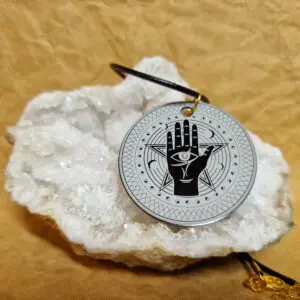 Le Talisman Sentinelle posé sur un cristal blanc présente une main protectrice avec un œil central entouré d’un motif sacré.