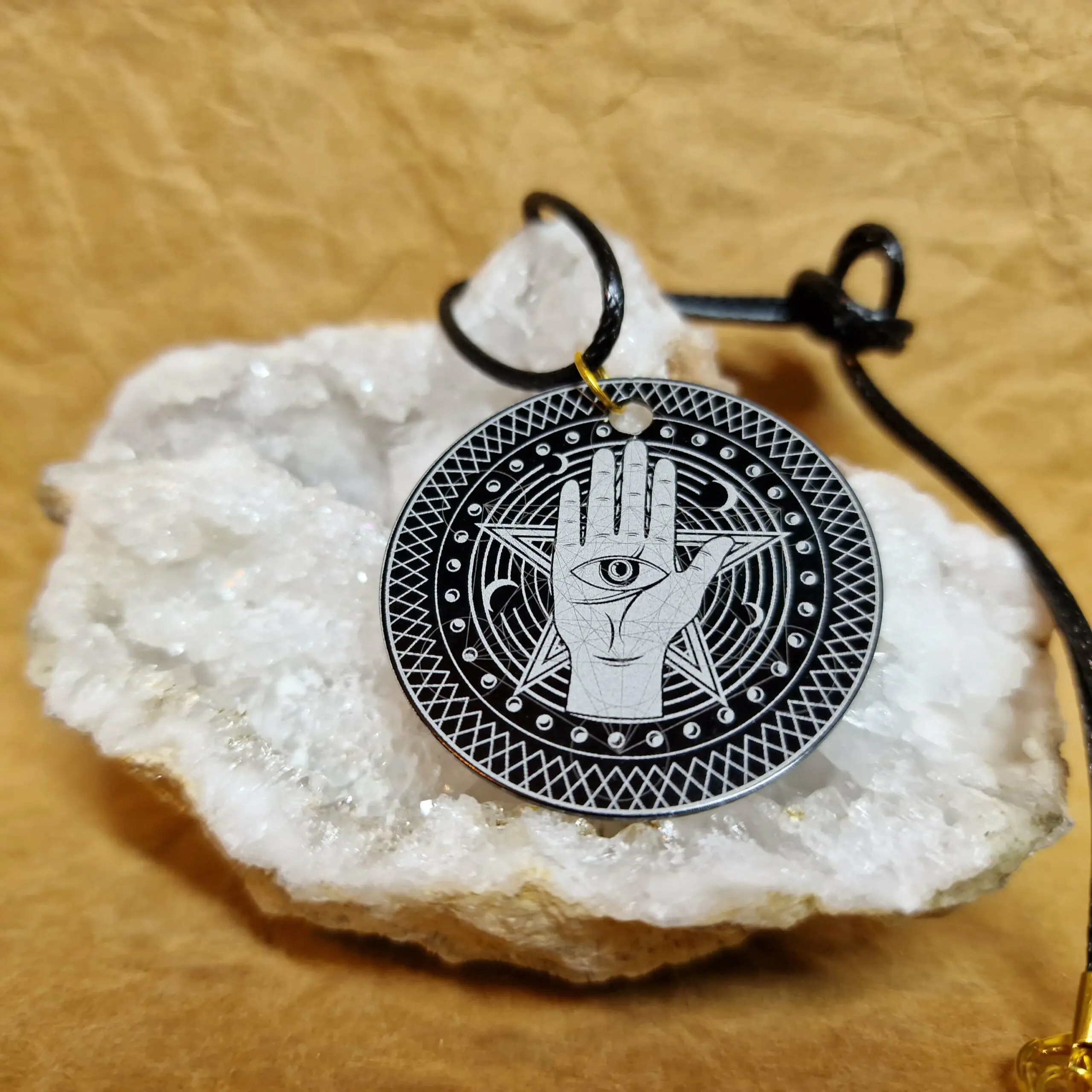 Le Talisman Sentinelle posé sur un cristal blanc présente une main protectrice avec un œil central entouré d’un motif sacré.