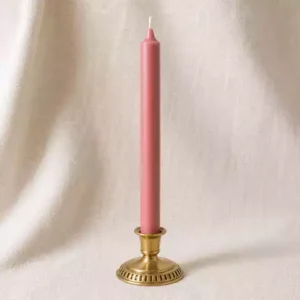 Bougie rose poudré fine dans un bougeoir en laiton sur fond lin crème avec lumière naturelle douce.