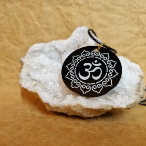 La médaille Le Talisman de la Vibration Ôm posée sur un cristal blanc montre le symbole Ôm entouré d’un motif floral sacré.