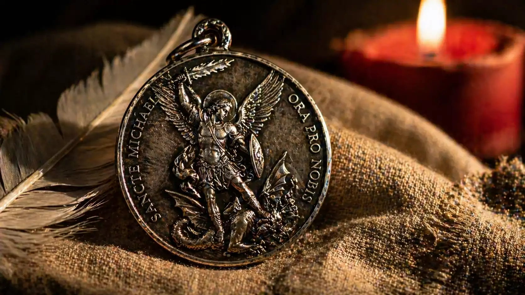 Médaille de Saint Michel Archange en argent vieilli terrassant le dragon, posée sur linge de lin avec plume blanche et bougie rouge.