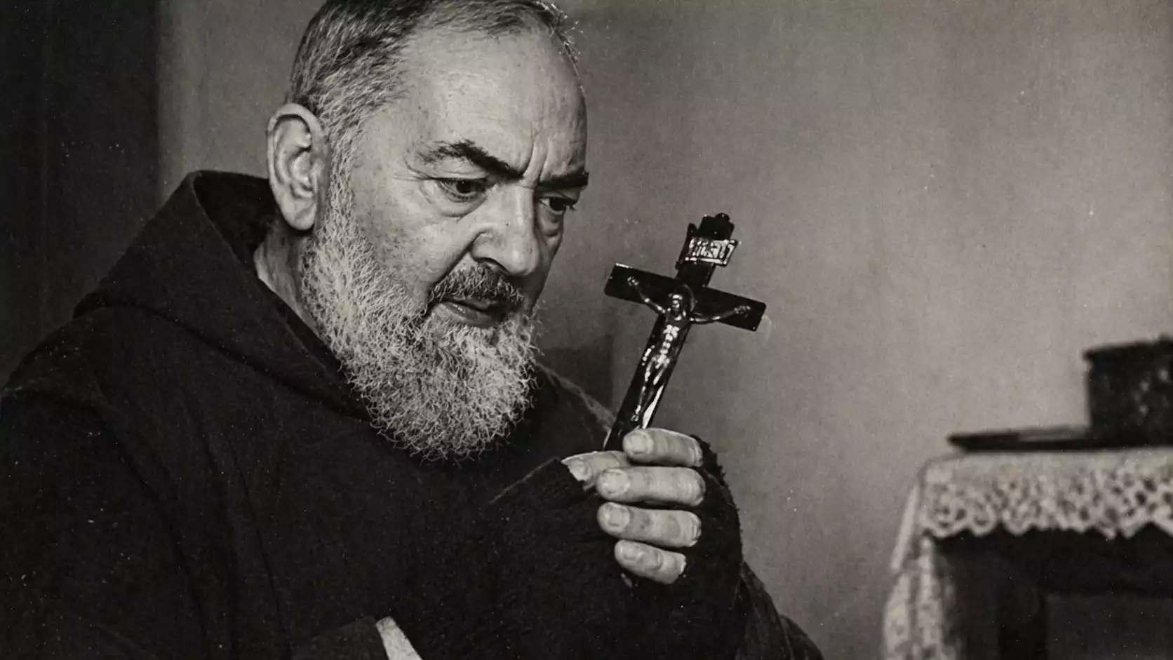 Padre Pio en habit capucin tenant un crucifix, avec mitaines noires sur les mains, dans une photographie noir et blanc au style archive.