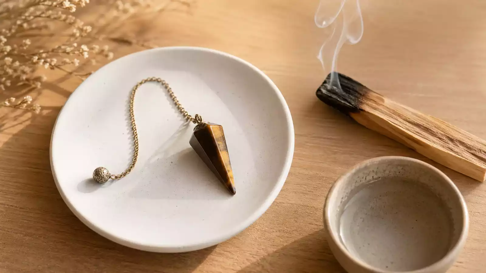 Pendule posé sur une assiette blanche avec palo santo fumant et bol d’eau claire sur fond bois chaleureux.