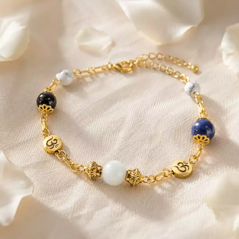 Bracelet énergétique doré avec pierres naturelles blanches, noires et bleues posé sur un linge en lin ivoire avec pétales blancs.