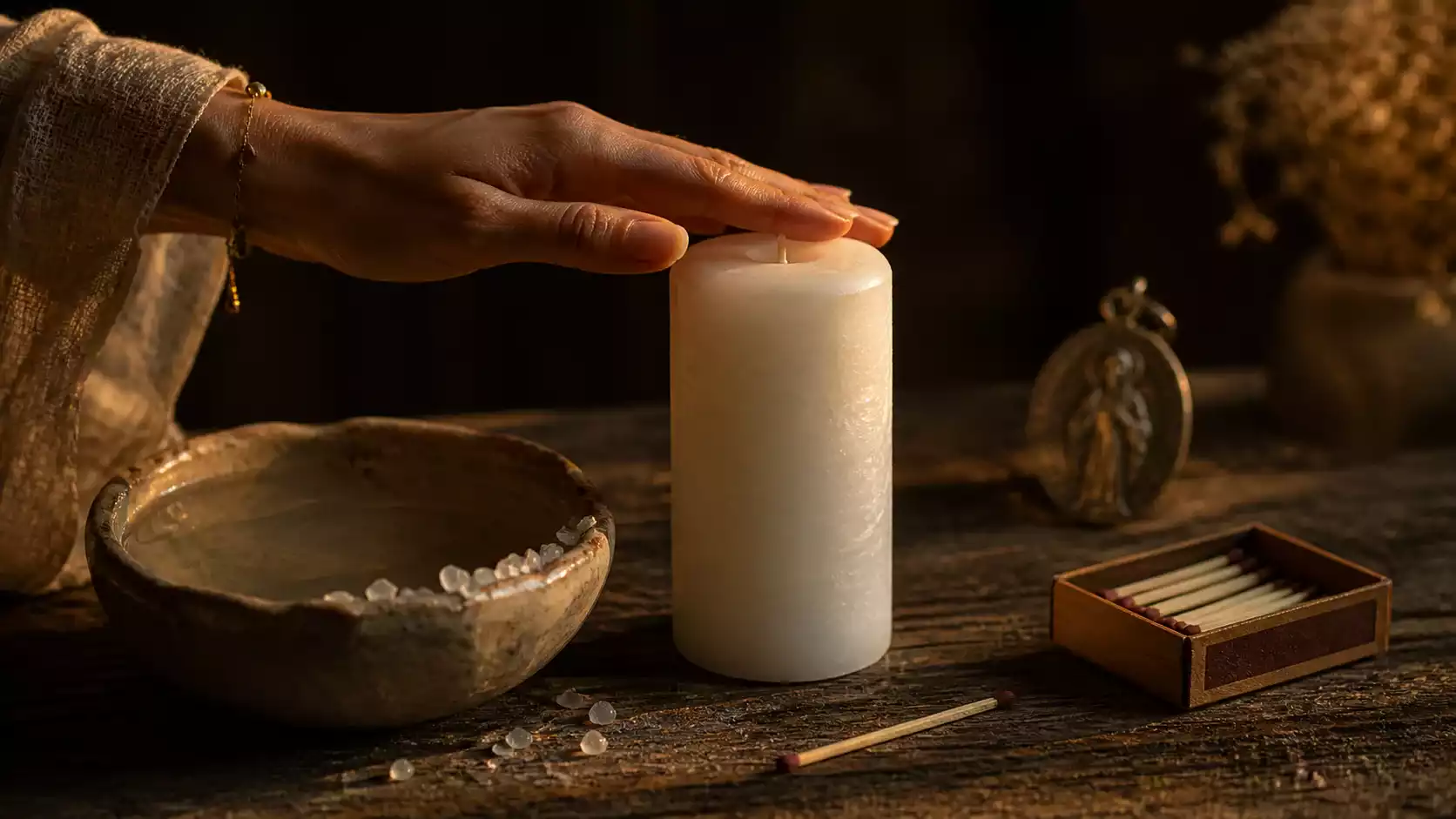 Main féminine effleurant une bougie blanche à côté d’une coupelle d’eau, de gros sel, d’allumettes et d’une médaille religieuse sur table en bois sombre.