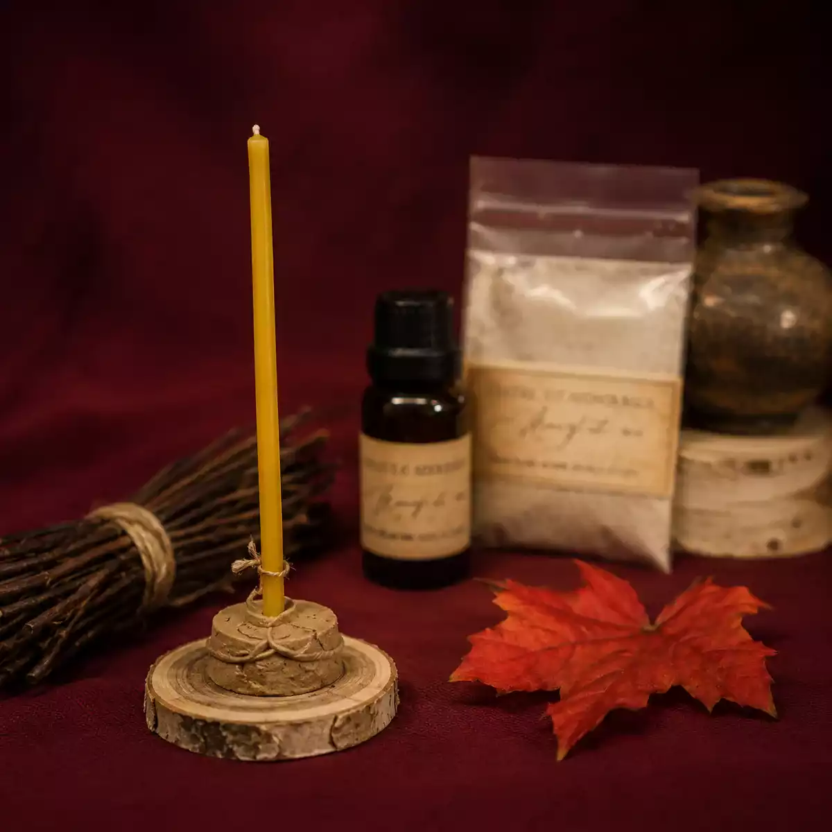 Mini-bougie rituelle couleur miel sur socle en bois avec flacon d’huile ambré, sachet de poudre, feuille d’érable rouge et décor automnal bordeaux.